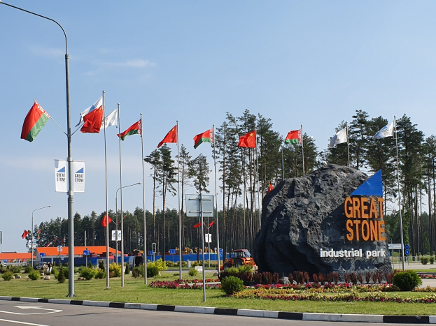 Großer Felsen mit der Aufschrift "Großer Stein Industriepark" vor grünem Gras, Blumen, Bäumen, fahnengeschmückten Laternenmasten, Gebäuden und einem blauen Himmel.