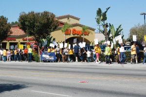 Eine Gruppe von Menschen mit Schildern marschiert eine Straße mit Bäumen, Büschen und Gebäuden mit Namensschildern entlang, mit Autos im Hintergrund und einem Banner im Hintergrund.