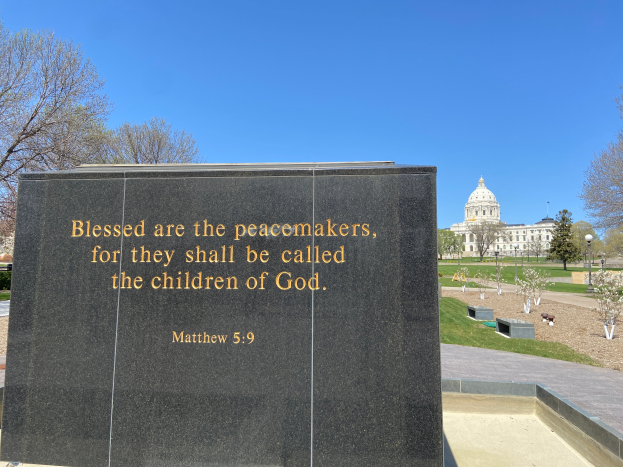 Ein Gedenkstein mit der Inschrift "Selig sind die Friedfertigen, denn sie sollen das Reich Gottes erben" umgeben von Bäumen, Pflanzen, Gras, Laternen und einem Gebäude unter einem klaren blauen Himmel.
