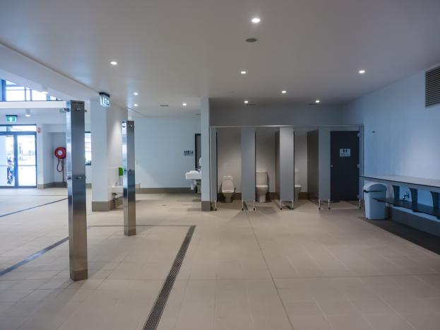 Großes öffentliches WC mit mehreren Toiletten, tragenden Säulen, einem Mülleimer, einer Tür, einem Schild, einem Feuerlöscher und Deckenleuchten.