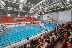 Großer Hallenbad mit vielen Menschen auf Stühlen drumherum und einigen im Wasser Stehenden, mit hoher Decke mit Lampen und Eisenstangen und Schildern mit Text im Hintergrund.