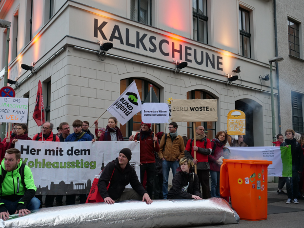 Eine Gruppe von Menschen mit Schildern und Plakaten steht vor einem Gebäude, zwei Personen sitzen im Vordergrund und ein Müllcontainer auf der rechten Seite während einer Protestaktion in Deutschland.