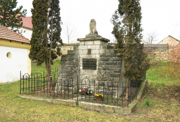 Ein steinerner Holocaust-Gedenkstein in einem grasbewachsenen Feld, umgeben von einem Metallzaun und geschmückt mit Blumenbouquets, mit Bäumen, Häusern und einer Wand im Hintergrund unter einem klaren blauen Himmel, Located in Sibiu, Romania.