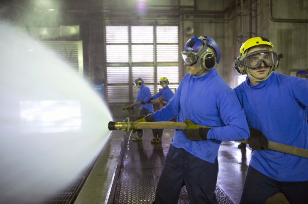 Gruppe von Männern in blauen Hemden und gelben Helmen bei der Arbeit an einer Maschine, wobei einer einen Schlauch hält und Wasser auf den Boden sprüht.