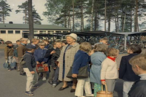 Eine Gruppe von Kindern steht vor einem Gebäude, einige halten Taschen und einer einen Korb, mit Fahrrädern, Schuppen, Bäumen und einem klaren blauen Himmel im Hintergrund, was an eine schwedische Schulszene der 1960er Jahre denken lässt.