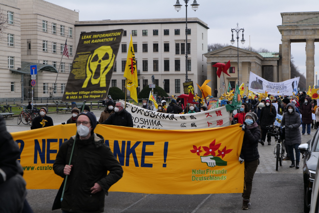 Eine große Gruppe von Menschen marschiert mit Schildern und Fahnen auf einer Straße während einer Demonstration gegen Atomkraft in Deutschland, mit Fahrzeugen auf der rechten Seite und Gebäuden, Laternenpfählen, Bäumen und einem Tor im Hintergrund.