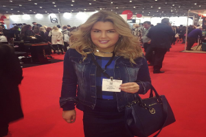 Frau auf einem roten Teppich bei einer Modenschau, die lächelt und eine Handtasche hält, mit Menschen, Lichtern und Schildern im Hintergrund.