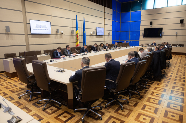 Große Gruppe von Menschen an einem langen Tisch in einem formellen Sitzungssetting mit EU-Flaggen, Fernsehern und Deckenleuchten in Brüssel.