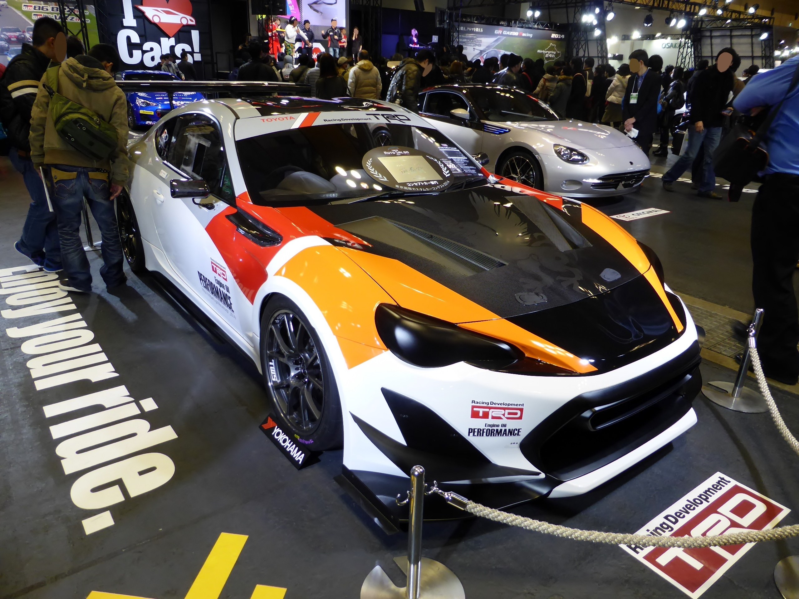 Eine Gruppe von Menschen um ein Rennfahrzeug auf der Tokyo Motor Show herumsteht, umgeben von Absperrpollern und Seilen, mit Informationsplakaten im Hintergrund unter beleuchteter Decke.