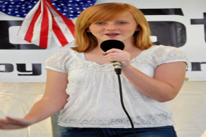 Eine Frau steht an einem Rednerpult und spricht in ein Mikrofon, mit einer USA-Flagge und einem Banner dahinter.