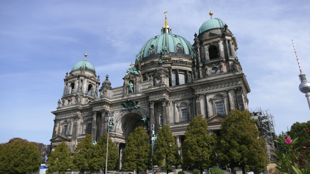 Große Kuppelkathedrale mit detaillierter Architektur, einschließlich Säulen, Bögen und Statuen, vor einem bewölkten Himmel, mit Passanten, Bäumen und einem Turm im Vordergrund.