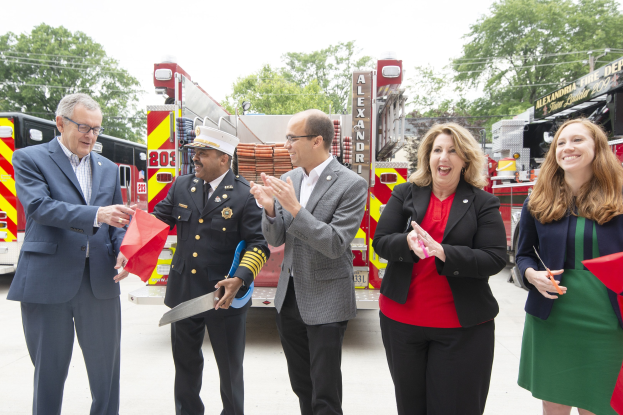 Gruppe von Menschen bei einer Eröffnungszeremonie der Alexandria Fire Department, die vor einem Feuerwehrauto klatschen und lächeln, während zwei eine Schere und ein rotes Band halten, Bäume und Drähte sind im Hintergrund unter einem klaren blauen Himmel zu sehen.