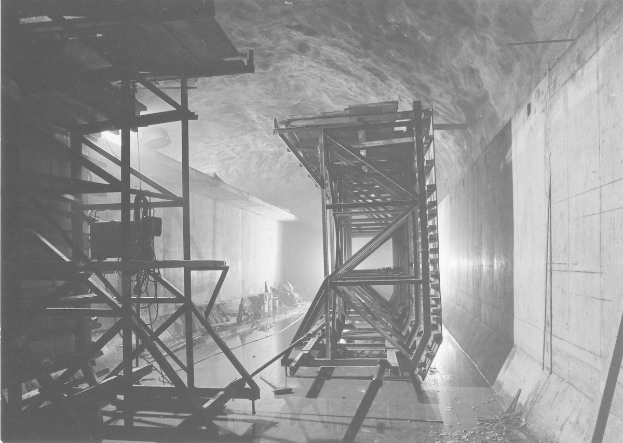 Schwarzes und weißes Foto eines Tunnels mit einer zentralen Metallstruktur, Metallstangen auf der linken Seite, einer Wand auf der rechten Seite und Text am unteren Rand.