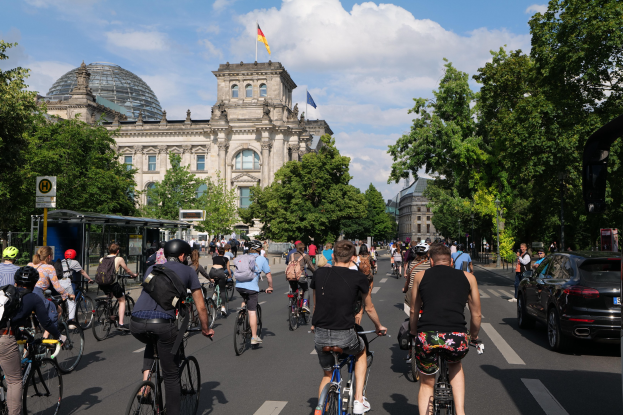 Gruppe von Menschen, die auf Fahrrädern eine von Bäumen gesäumte Straße in Berlin, Deutschland, entlangfahren, mit Gebäuden und einer Bushaltestelle im Bild, unter einem bewölkten Himmel und einer Flagge, die an einem Gebäude weht.
