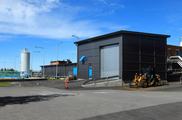 Großes Industriegebäude mit Bulldozer davor, umgeben von Straßeninfrastruktur und Fahrzeugen, unter einem bewölkten Himmel.