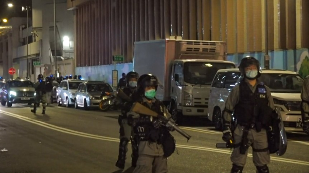 Polizeibeamte in Helmen, Handschuhen und Gesichtsmasken stehen nachts auf einer Straße, einige halten Waffen, mit Fahrzeugen, Laternenpfählen, Schildern und Gebäuden im Hintergrund.