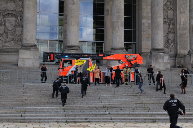 Menschen gehen Treppe vor einem Gebäude mit Glasfenstern, Säulen und Verzierungen herunter, neben einem geparkten Fahrzeug und einem Metallzaun, einige tragen Mützen und einer hält ein Objekt.