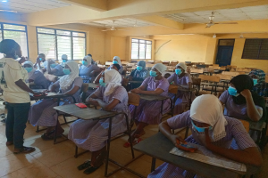 Ein Klassenzimmer in Nigeria mit Schülern, die Gesichtsmasken tragen und auf Bänken und Stehplätzen sitzen, mit Büchern und Gegenständen auf den Tischen, Fenstern, einer Tür, einer Wand, einer Decke mit Lampen und Ventilatoren im Hintergrund.