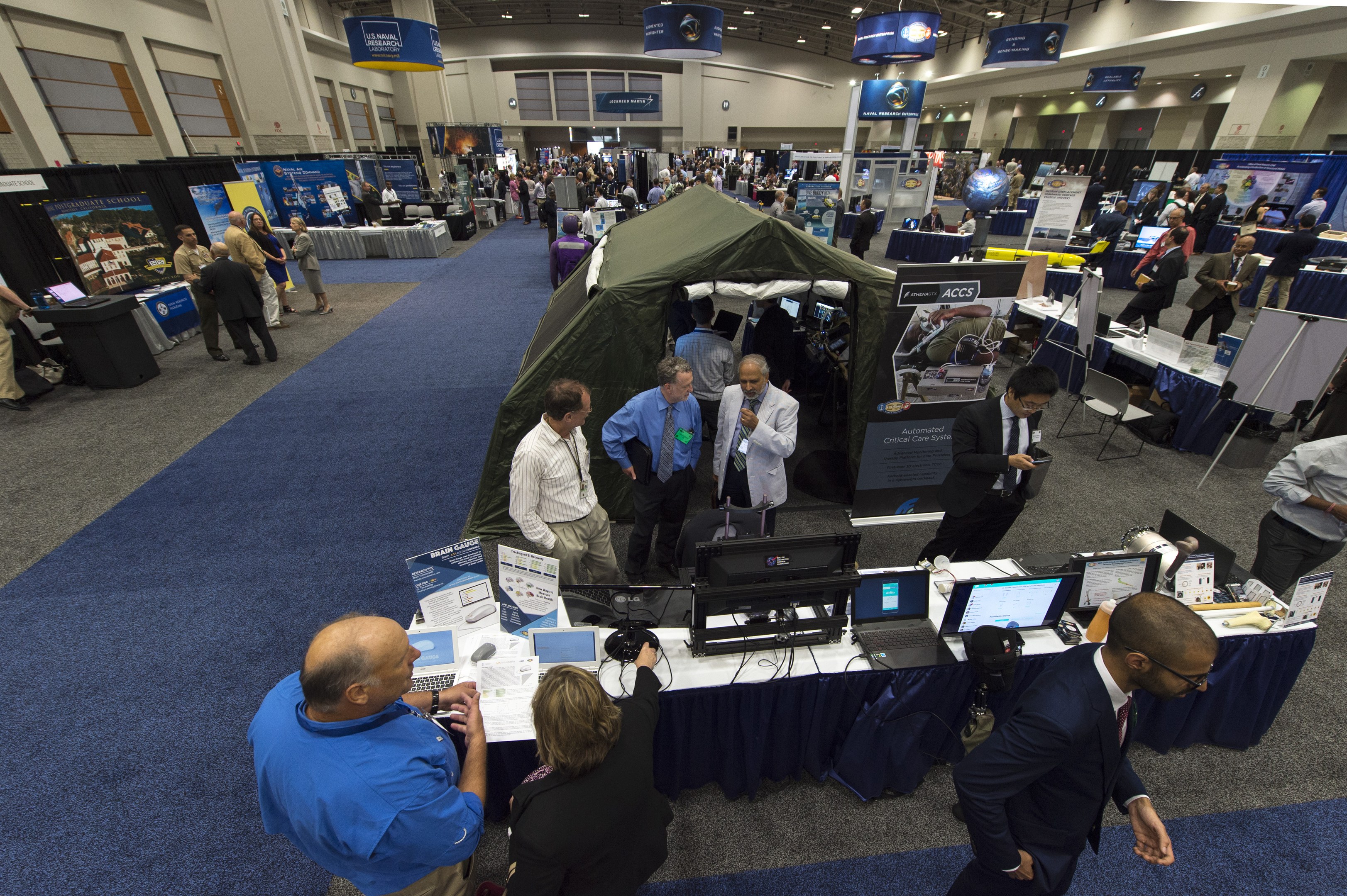 Eine große Gruppe von Menschen auf einer Karriere-Messe steht um Tische mit Laptops, Monitoren und Papieren, mit einem Zelt, Tafeln, Deckenleuchten und Fenstern im Hintergrund.