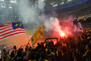 Eine große Menge Menschen in einem Stadion mit Bengalos, Fahnen und einer Kamera, mit Rauch und Schildern mit der Aufschrift "Malaysia vs Malaysia Live Streaming" im Hintergrund.