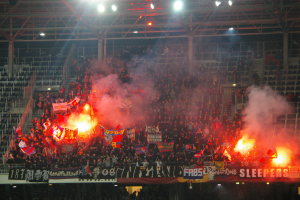 Eine große Menge in einem Stadion hält Fahnen und Banner, mit Leuchtraketen, die Rauch erzeugen, unter einem Dach mit Deckenleuchten und Metallrahmen.