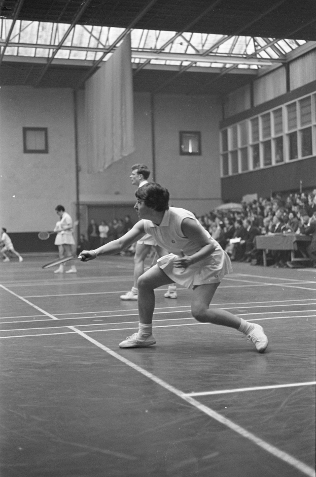 Schwarzes und weißes Foto von zwei Frauen, die Badminton auf einem Court spielen, mit Rackets in den Händen, umgeben von Zuschauern auf der rechten Seite und einer Wand mit Fenstern im Hintergrund.