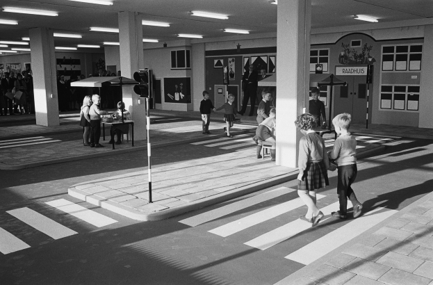 Schwarz-weiß-Foto von Kindern, die eine Straße an einer Ampel überqueren, mit Fußgängern in der Nähe, einem Verkehrssignalmast im Vordergrund und strukturellen Säulen mit Deckenleuchten im Hintergrund.