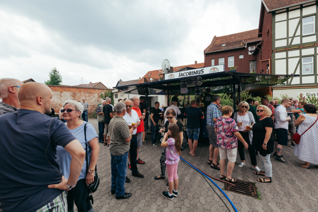 Gruppe von Menschen auf einem Outdoor-Bierfest vor einem Gebäude mit Fenstern, Bäumen und einem Schuppen mit einem Namensschild im Hintergrund.