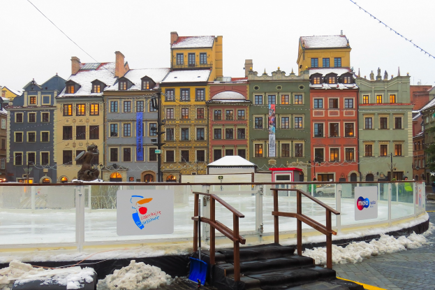 Eisbahn in einem Stadtplatz umgeben von Gebäuden, Treppen, Laternen, Bäumen und schneebedeckter Erde unter einem Himmel.