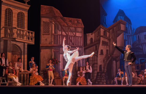 Zwei Menschen tanzen auf einer Bühne mit einem projizierten Stadtpanorama-Hintergrund im Royal Opera House in London, während das Publikum zuschaut.