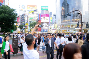 Ein Mann mit einem Handy geht auf einem Weg mit anderen Menschen, umgeben von Bäumen, Pfählen, Ampeln, Gebäuden und Schildern.
