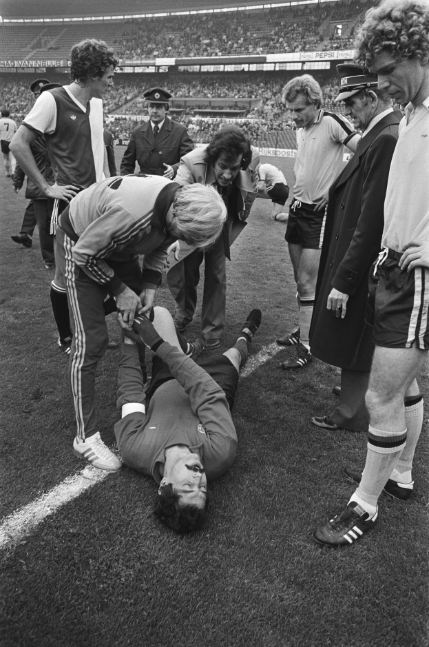 Schwarzes Bild eines Fußballspielers, der auf dem Boden liegt und von einer Gruppe Männer umgeben ist, während eine Menge in einem Stadion zusieht.