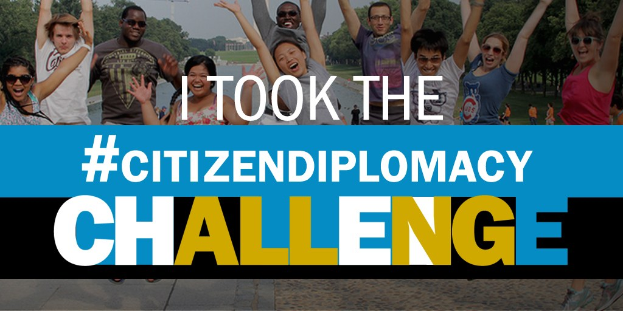 Eine Gruppe von Menschen, die vor einem Schild mit der Aufschrift "Ich habe die Staatsbürgerschaft angenommen" posieren und lächeln, mit Bäumen, Gebäuden, Kränen und einem klaren blauen Himmel im Hintergrund.