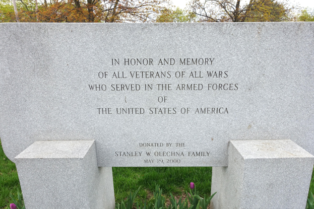 Gedenkstein mit einer Inschrift für US-Militärveteranen, umgeben von Pflanzen, Blumen, Gras, Bäumen und einem bewölkten Himmel.