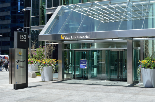 Moderner gläserner Sun Life Financial Tower in Toronto mit einer Leuchttafel, Topfpflanzen mit Blüten, Passanten, einer Straßenlaterne, einer Leuchttafel, Fahrzeugen auf der Straße und einem bewölkten Himmel.