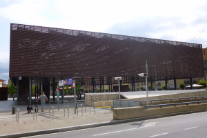 Großes modernes Gebäude mit zahlreichen Fenstern, das als neuer Eingang ins Stadtzentrum dient, flankiert von Straßeninfrastruktur und Fußgängern.