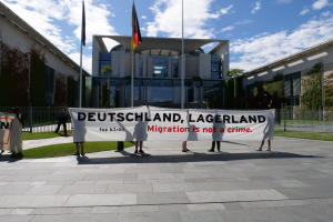 Gruppe von Menschen mit einem Banner 'Deutschland, Lagerland Migration ist kein Verbrechen' vor einem umzäunten Gebäude mit Bäumen und Fahnen.