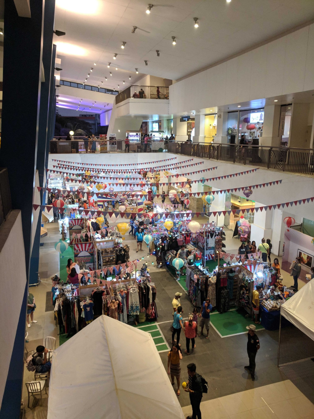 Ein volles Festivitätseinkaufszentrum mit Ständen, hängenden Kleidern, Dekorationsfahnen, Ballons, sitzenden und stehenden Menschen, Geländern, Säulen und Deckenleuchten, das einem Weihnachtsmarkt im Mall of Asia ähnelt.