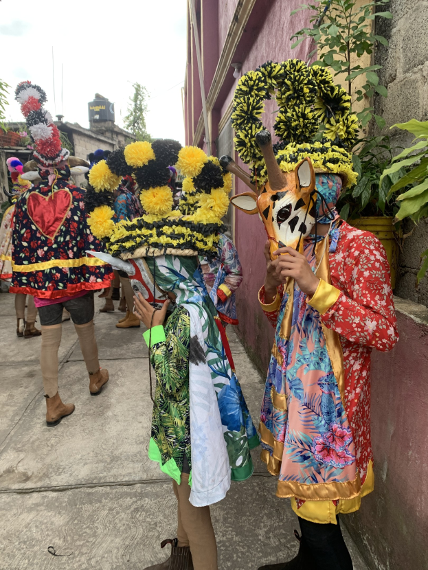 Eine Gruppe von Menschen in bunten Kostümen steht vor einem Gebäude während eines Faschingsumzugs, mit verschiedenen Gegenständen in den Händen, Bäumen, Gebäuden und einem klaren blauen Himmel im Hintergrund.