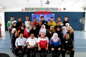 Eine Menschenmenge, die fröhlich vor einer Bühne posiert und ein Schild mit Text an der Wand im Hintergrund, ein Tischventilator rechts, ein Podium links und ein Projektor oben im Bild.