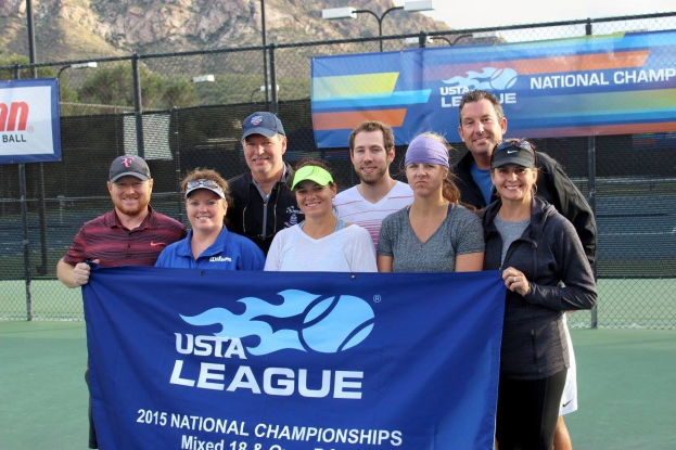 Gruppe von Menschen auf einem Tennisplatz mit einem "2015 USTA National Championships"-Schild, einem Zaun, Bannern, Laternen, Bäumen, Bergen und einem klaren blauen Himmel im Hintergrund.
