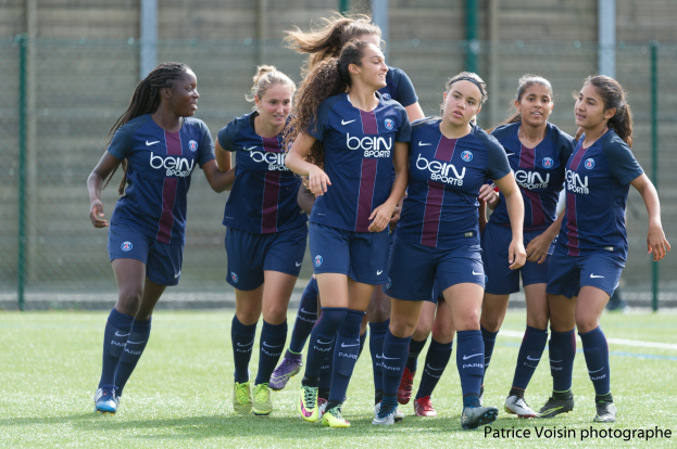 Gruppe junger Frauen beim Fussballspielen auf einem Rasenplatz mit Maschendrahtzaun und einer Wand im Hintergrund, beschriftet mit "Paris Saint-Germain Frauenfussball" in der rechten unteren Ecke.