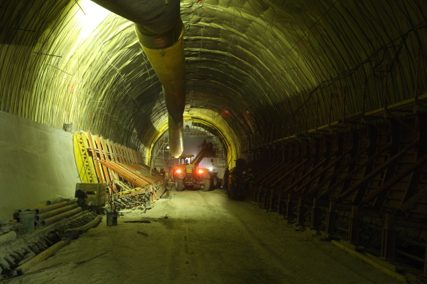 Baustelle mit einem großen Tunnel, Fahrzeugen, verstreuten Holzobjekten, Rohren, einer Wand auf der linken Seite, Überkopfrohren und beleuchteten Tunnel-Lampen.