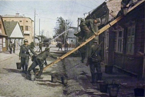 Gruppe von Weltkriegsoldaten deutscher Wehrmacht in militärischer Uniform auf einer Straße neben einem Gebäude stehend, mit Helmen und hölzernen Gegenständen bewaffnet, mit Eimern auf dem Boden und Gebäuden, Bäumen, Pfählen, Drähten und einem klaren Himmel im Hintergrund.