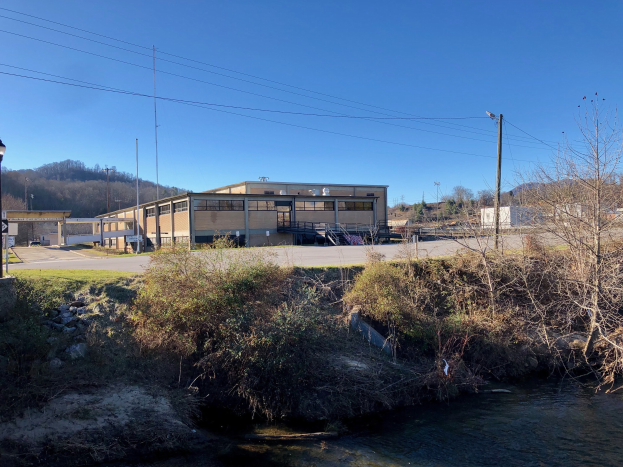Grüne Wiese mit einem Fluss, Bäumen, Pflanzen, Gras, Steinen, Straßenlaternen, Strommasten, Stromkabeln, Hügeln und einem klaren blauen Himmel, mit einem Gebäude an der geplanten Stelle einer Wasseraufbereitungsanlage.