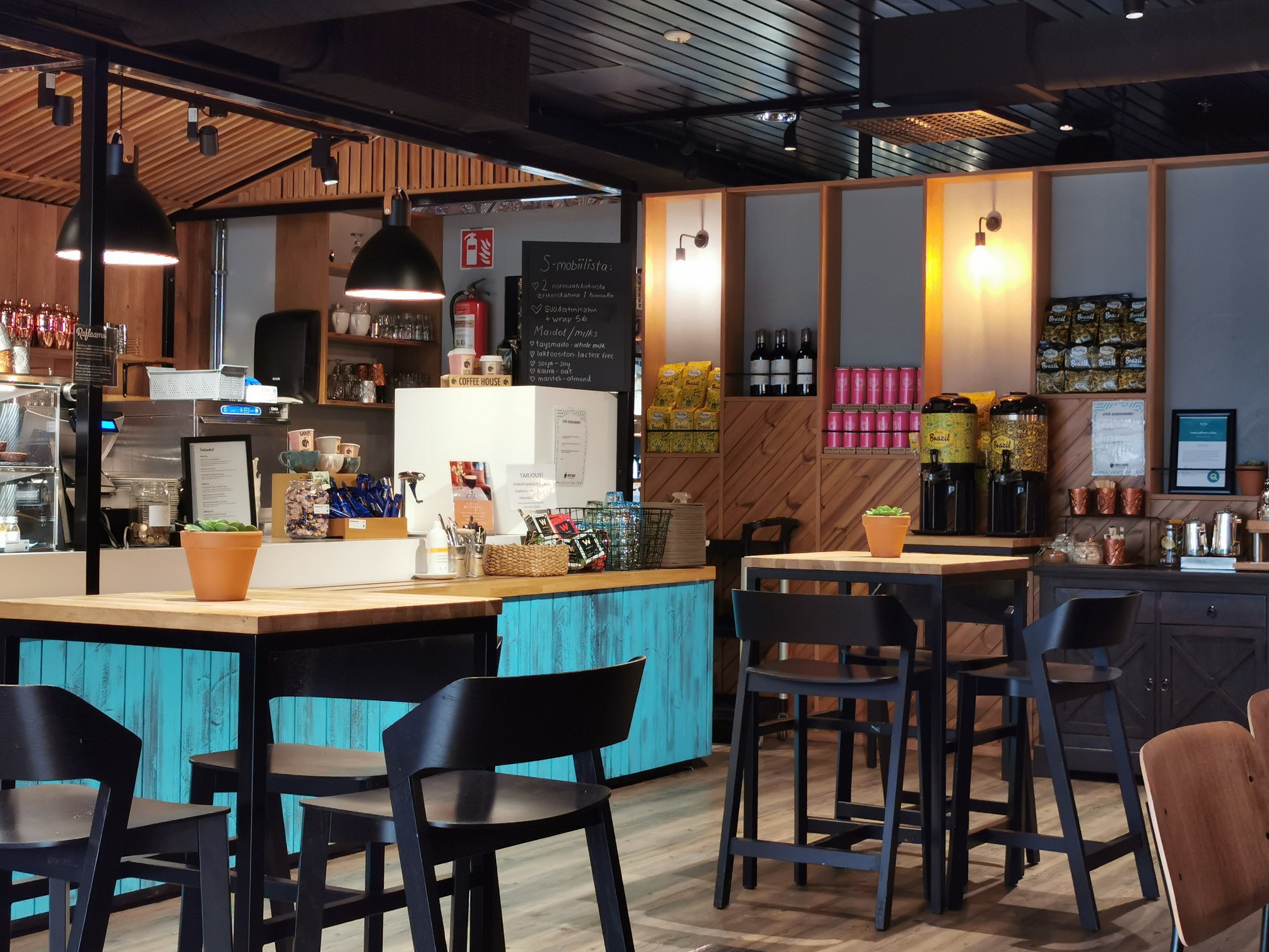 Innenraum eines Restaurants mit Holztischen, Stühlen, einer Topfpflanze auf einem Tisch, einer Schautafel, einem Kühlschrank, einem Feuerlöscher, Regalen mit Flaschen, Schränken mit Gegenständen, einem Foto auf der Wand und Deckenleuchten.