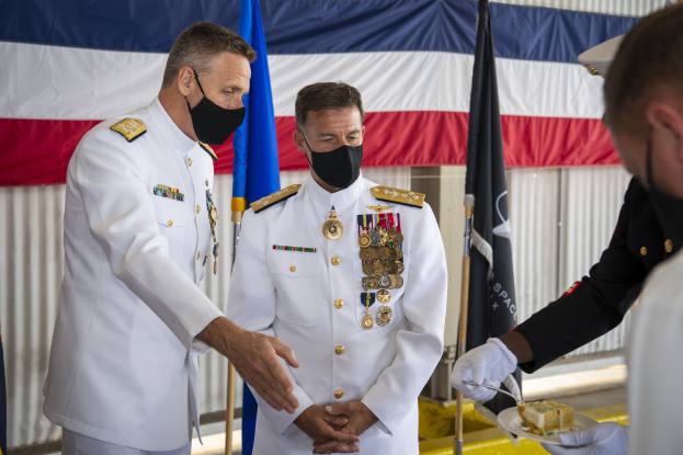Zwei Männer in weißen Uniformen und Masken stehen neben Flaggen und einem Vorhang, wobei einer ein Tablett mit einem Lebensmittel hält, das den 50. Jahrestag der US-Navy feiert.