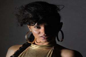 Eine Frau in einem goldenen Kleid und großen Ohrringen posiert selbstbewusst vor einer Wand, ihr glatter Bob und natürliches Make-up unterstreichen ihren entschlossenen Gesichtsausdruck, während sie direkt in die Kamera blickt.