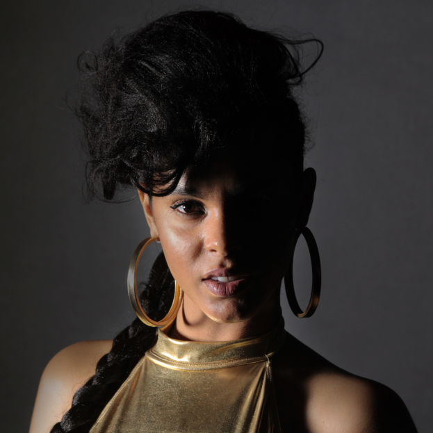 Eine Frau in einem goldenen Kleid und großen Ohrringen posiert selbstbewusst vor einer Wand, ihr glatter Bob und natürliches Make-up unterstreichen ihren entschlossenen Gesichtsausdruck, während sie direkt in die Kamera blickt.