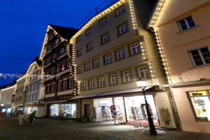 Eine Kopfsteinpflasterstraße in Rothenburg ob der Tauber, Deutschland, geschmückt mit festlichen Weihnachtslichtern, mit Menschen, die spazieren gehen und einem Fahrrad in der Nähe, flankiert von Gebäuden mit Fenstern unter einem sichtbaren Himmel.